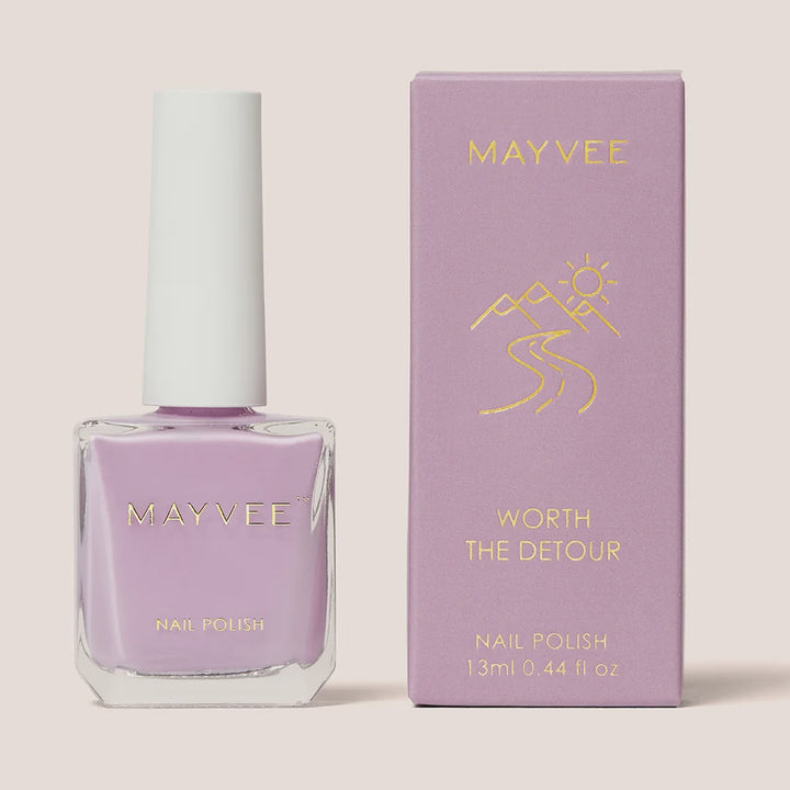A bottle of MAYVEE nail polish in a light lavender shade stands next to its matching lavender box with gold foil lettering, set against a solid light beige background.