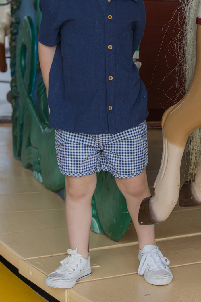 Child wearing a navy Milky shirt, checkered shorts, and white sneakers standing on a tiled floor.