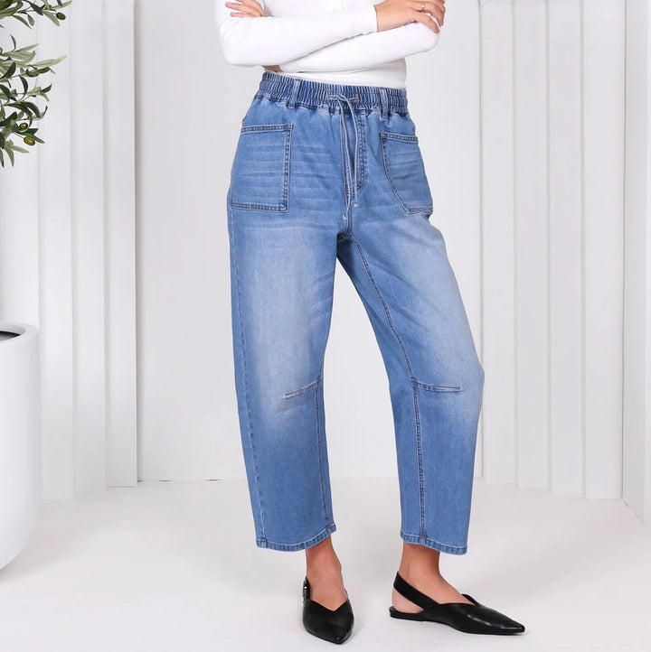 Woman wearing a white top and blue jeans standing in a minimalistic room with a plant.