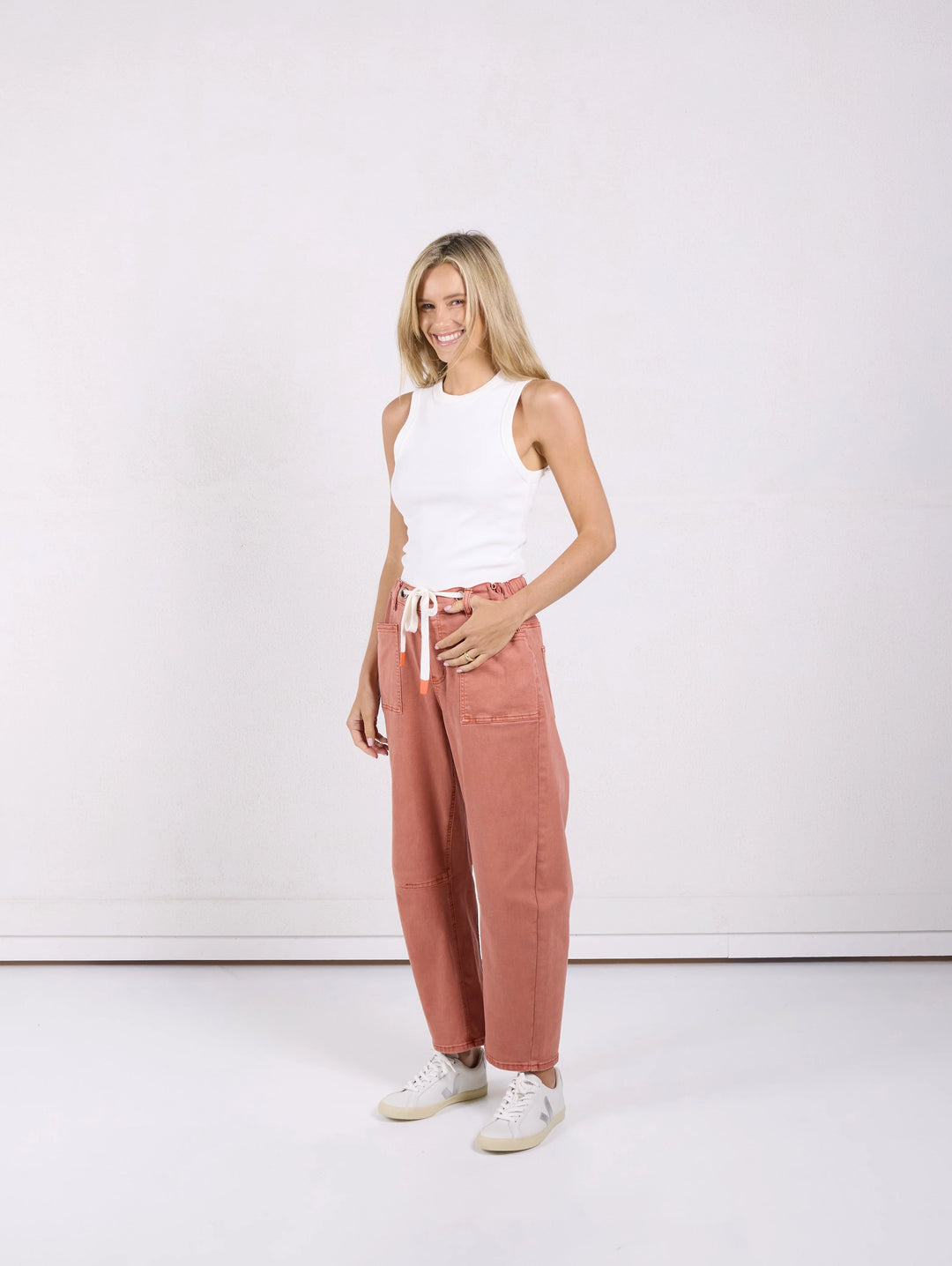 A woman modeling a pair of wide-leg, dusty rose pants with a white drawstring, paired with a white sleeveless top and white sneakers, standing against a plain white studio background.
