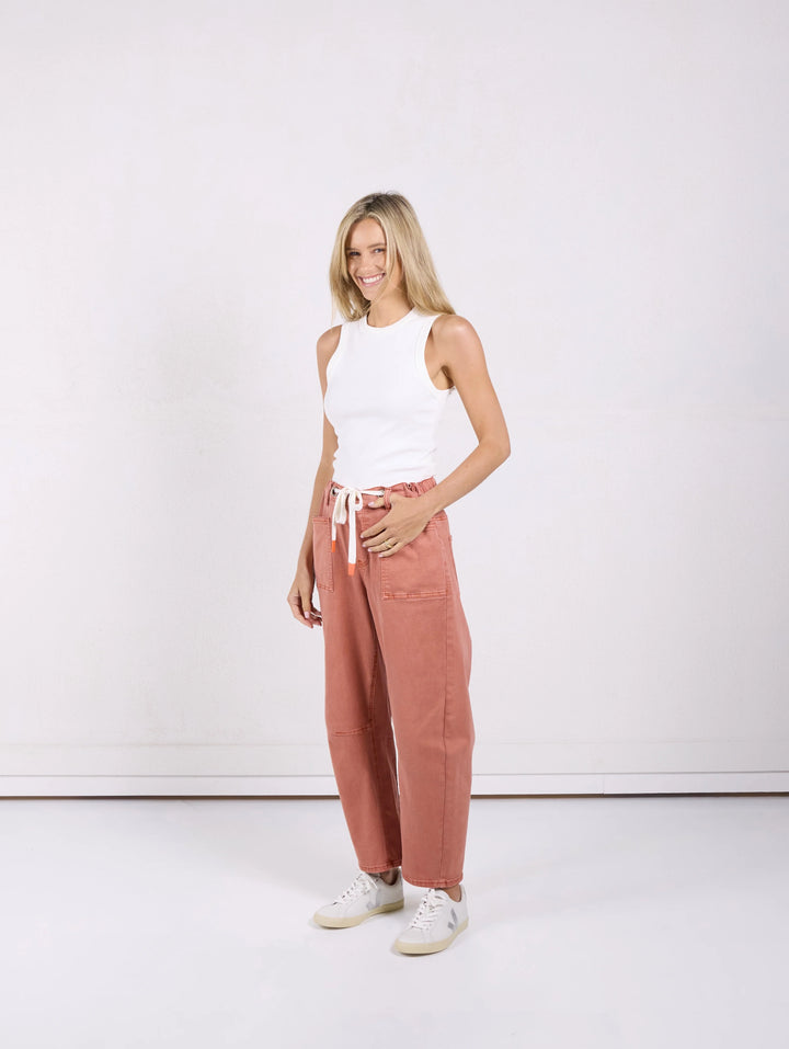 A woman modeling a pair of wide-leg, dusty rose pants with a white drawstring, paired with a white sleeveless top and white sneakers, standing against a plain white studio background.