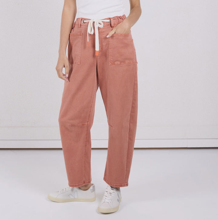A woman modeling a pair of dusty rose wide-leg pants with a white drawstring, paired with a white sleeveless top and white sneakers, standing against a white studio background.