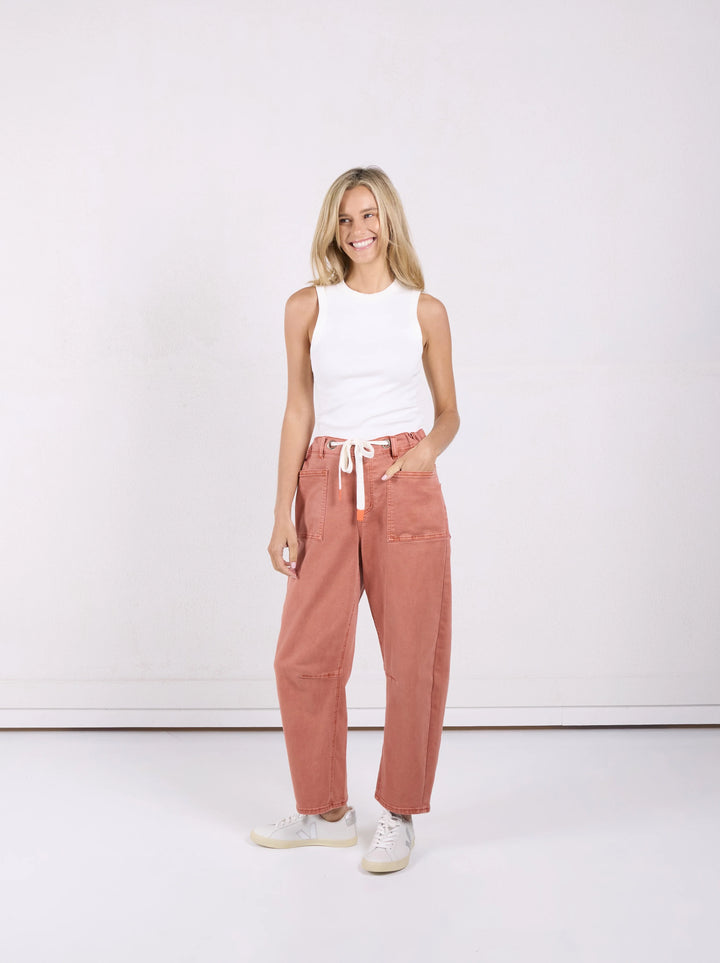 A woman modeling a pair of dusty rose wide-leg pants with a white drawstring, paired with a white sleeveless top and white sneakers, standing against a white studio background.