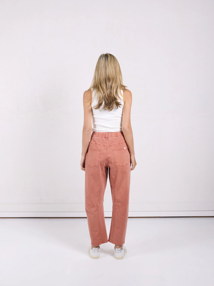 A woman viewed from behind wearing a white tank top and dusty rose wide-leg corduroy pants, standing against a plain white studio wall.