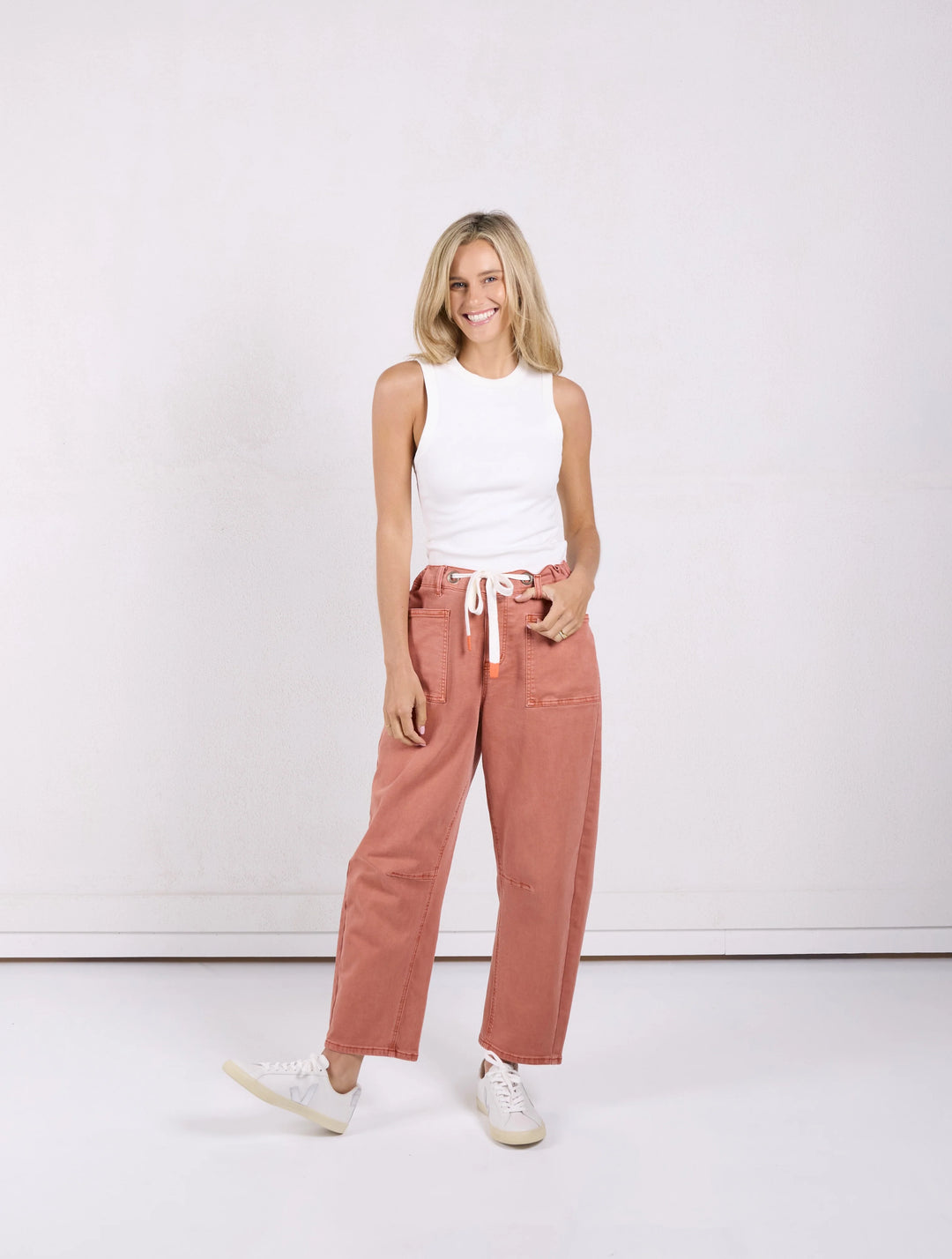 A woman modeling a pair of wide-leg, cropped wide-leg pants in a dusty rose color, paired with a white ribbed tank top and white sneakers, standing against a plain white studio background.