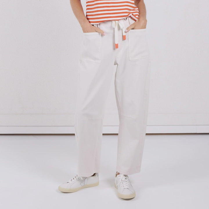 A woman modeling a short-sleeve orange and white striped knit top with white drawstring pants and white sneakers, standing against a plain white studio background.