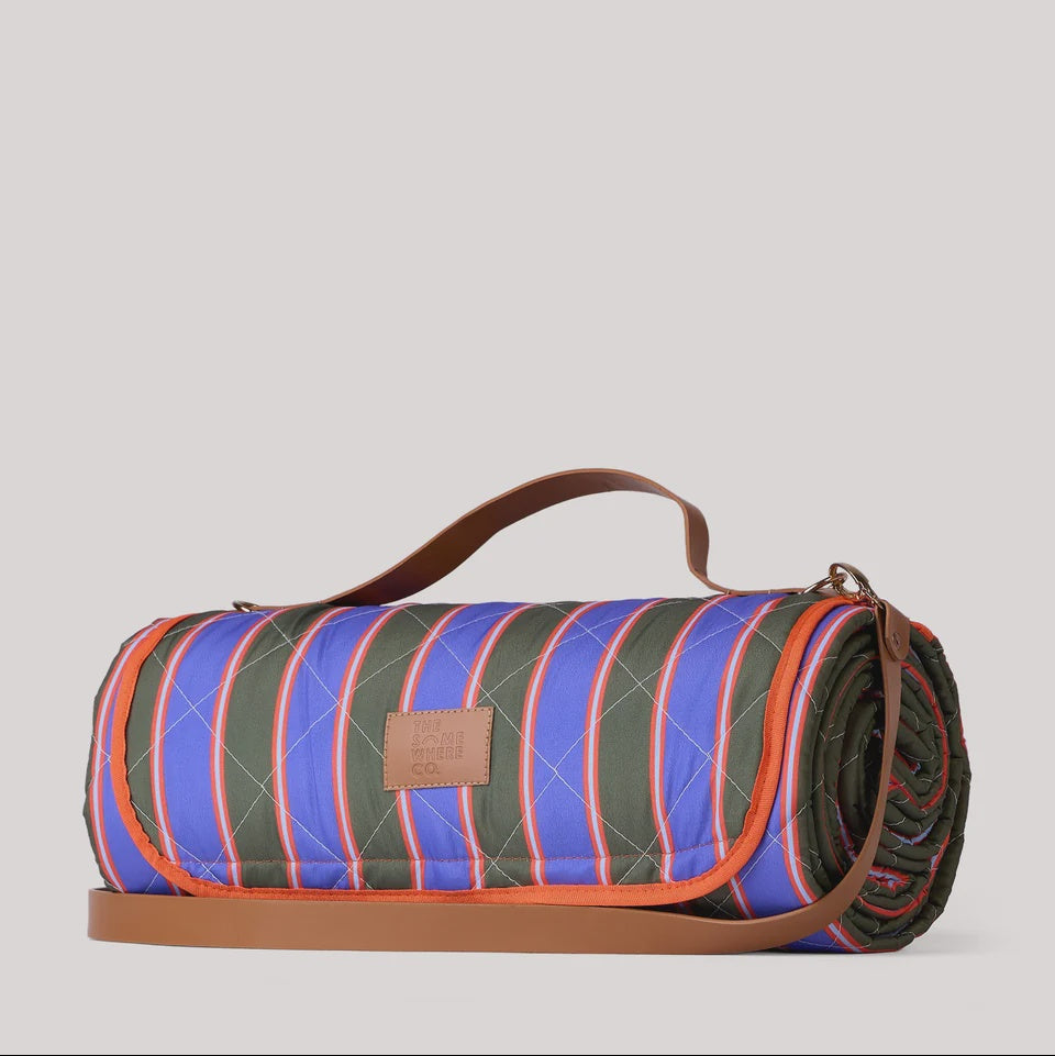 A rolled-up quilted picnic blanket with a vibrant blue, green, and orange striped pattern, featuring brown leather handles and trim, set against a solid light gray background.