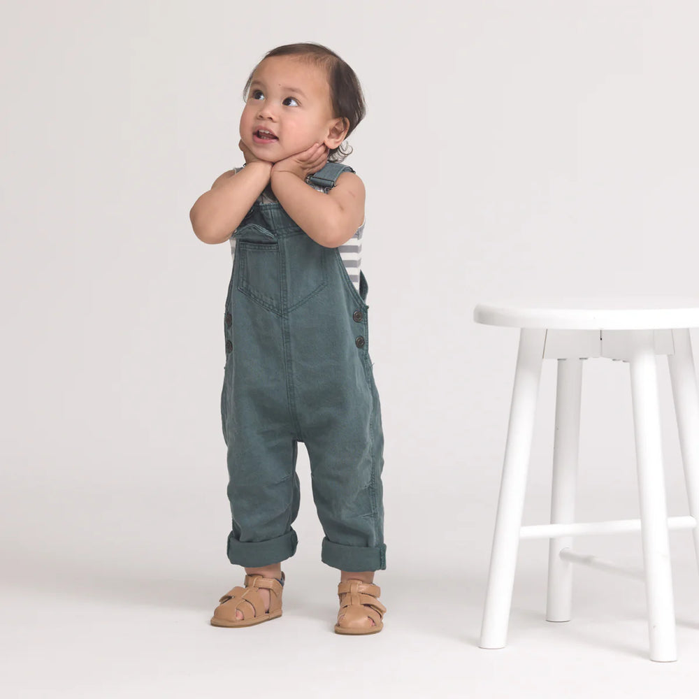Child wearing green overalls standing next to a white stool on a plain background