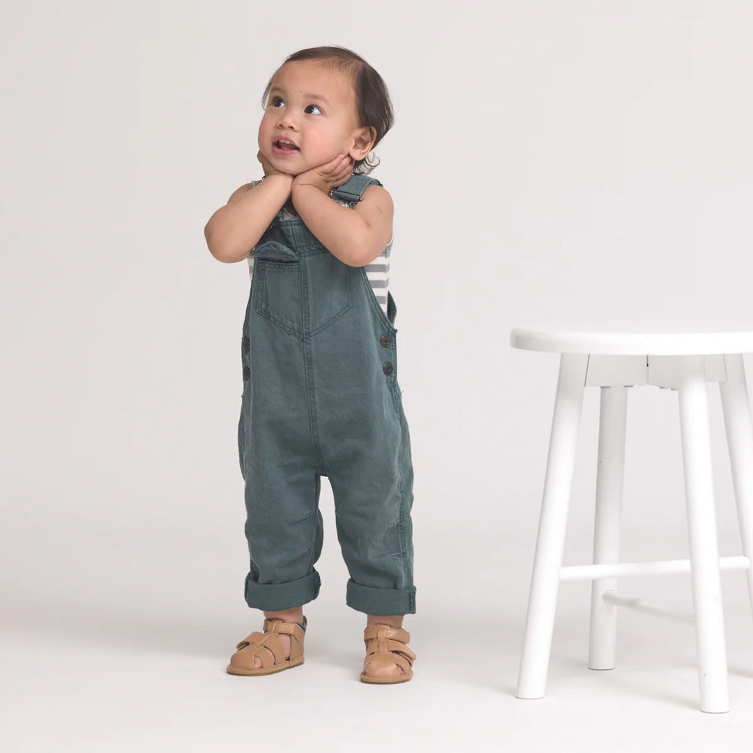 Child wearing green overalls standing next to a white stool on a plain background