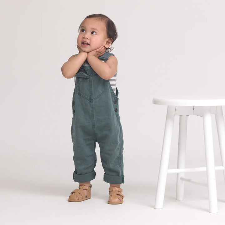 Child wearing green overalls standing next to a white stool on a plain background