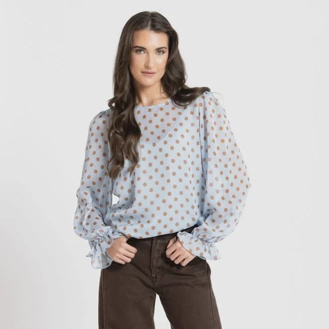A woman modeling a light blue long-sleeve blouse with orange polka dots and ruffled cuffs, paired with dark brown wide-leg trousers and black pointed flats, against a light gray studio background.