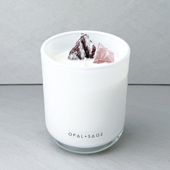 A white Opal + Sage scented candle in a clear glass jar, decorated with two crystals on top, one dark brown and one pink, set against a neutral gray background.