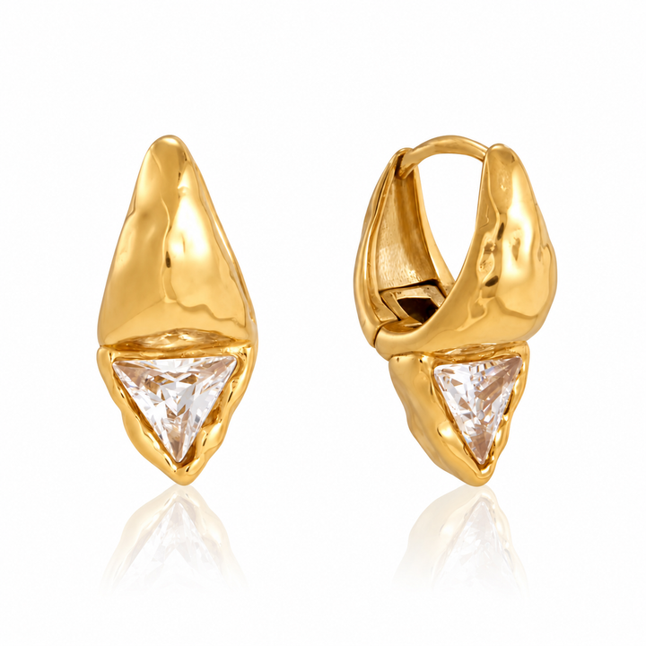 A pair of gold huggie hoop earrings with a textured, organic triangular gold setting and a clear faceted triangle gemstone at the bottom, shown on a white background with a soft reflection.