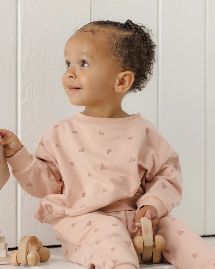 A toddler wearing a light pink long-sleeve sweatshirt and matching sweatpants set with a small heart pattern, sitting on a white wooden floor next to wooden toy cars.