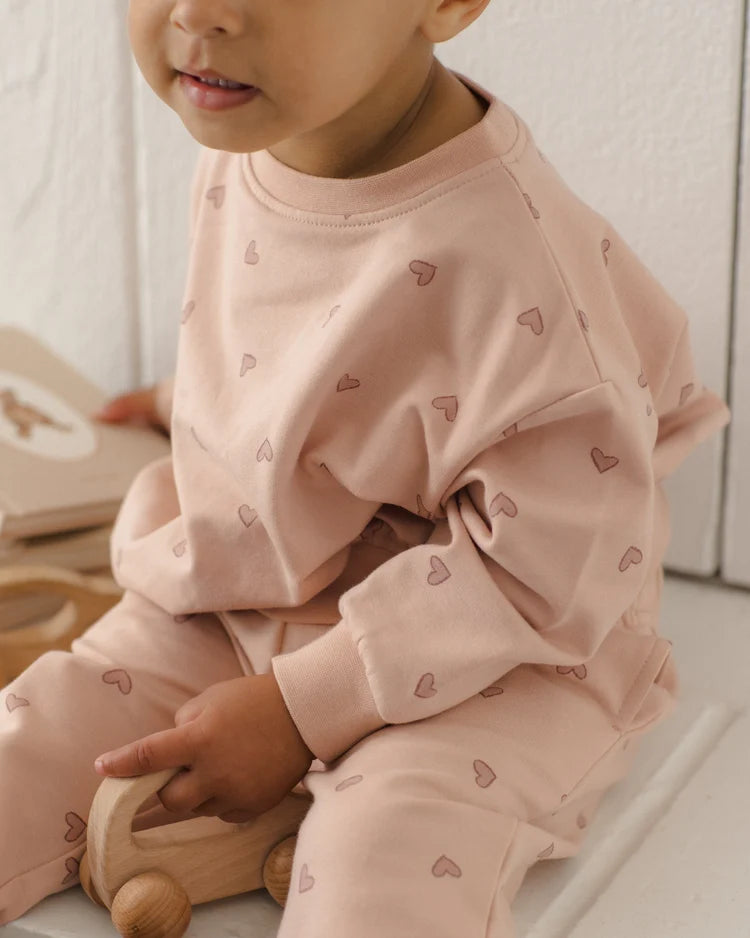 A baby wearing a light pink two-piece pajama set with a subtle heart pattern, sitting next to a wooden toy car.