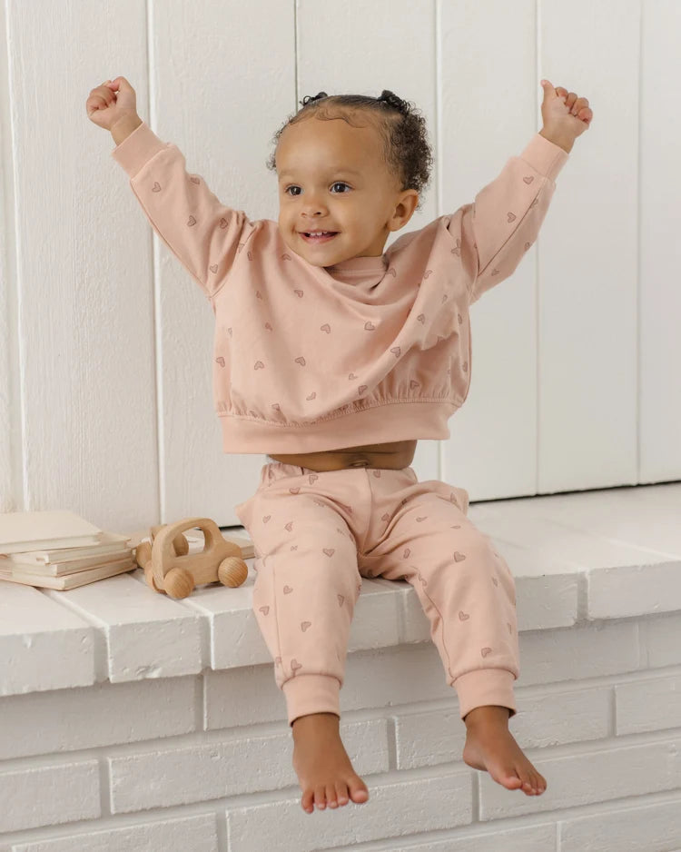 A toddler girl wearing a matching dusty pink two-piece loungewear set with a subtle heart pattern, sitting on a white brick ledge with her arms raised in excitement.