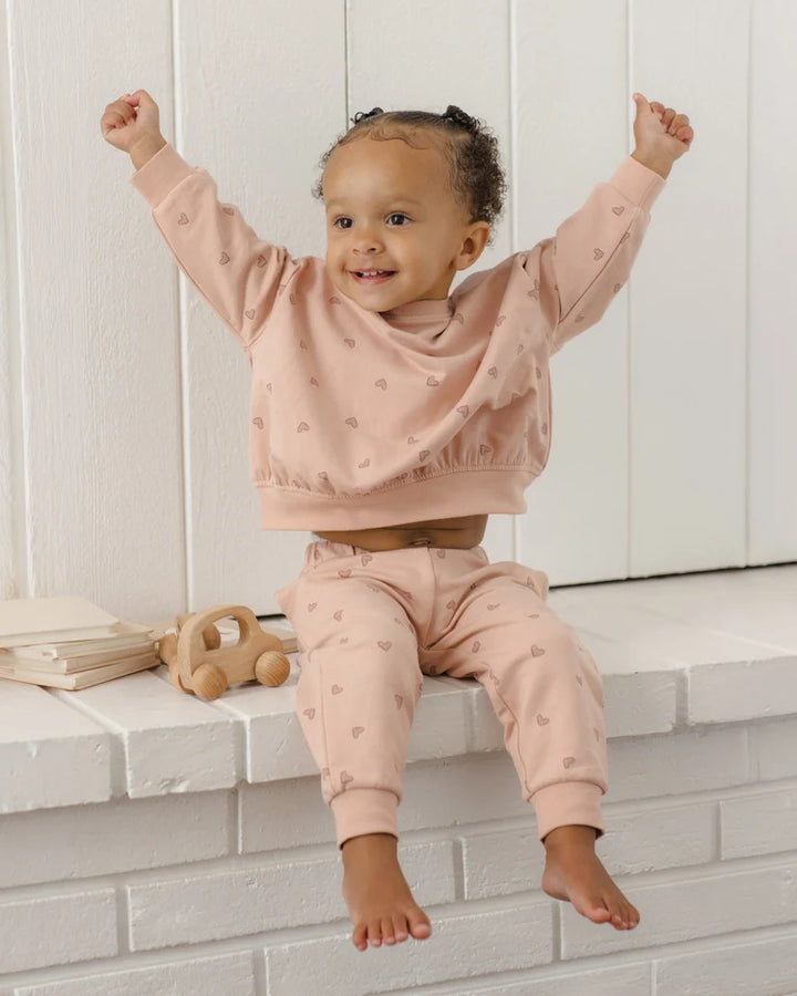 A toddler girl wearing a matching dusty pink two-piece loungewear set with a subtle heart pattern, sitting on a white brick ledge with her arms raised in excitement.