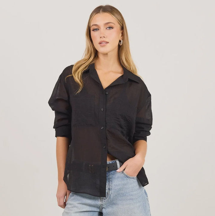 A woman modeling a black sheer long-sleeve button-up shirt and light-wash blue denim culottes, paired with black heeled thong sandals, against a plain off-white studio background.