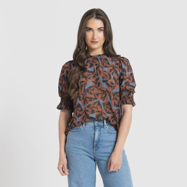 A woman modeling a brown and blue patterned blouse with puff sleeves, paired with wide-leg light blue jeans and black pointed flats, against a light gray studio background.