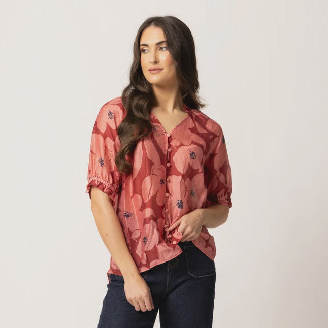 A woman modeling a short-sleeve button-up blouse with a dusty rose and red abstract floral pattern, paired with dark wide-leg jeans and brown heeled sandals, against a plain light beige studio background.