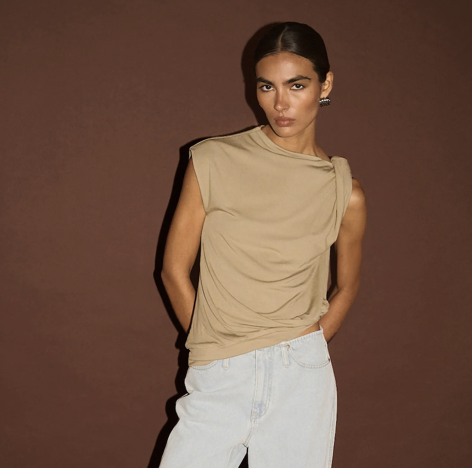 Woman wearing a beige sleeveless top and light blue jeans against a brown background