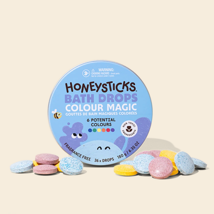A round tin of Honeysticks Bath Drops Colour Magic bath drops sits on a light beige background with several loose pastel-colored drops scattered around the base. The tin is light blue with purple cartoon graphics and text indicating 6 potential colours and 36 drops.