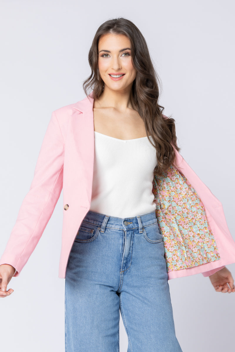 Woman wearing a pink seeking lola blazer over a floral shirt with blue jeans on a light gray background