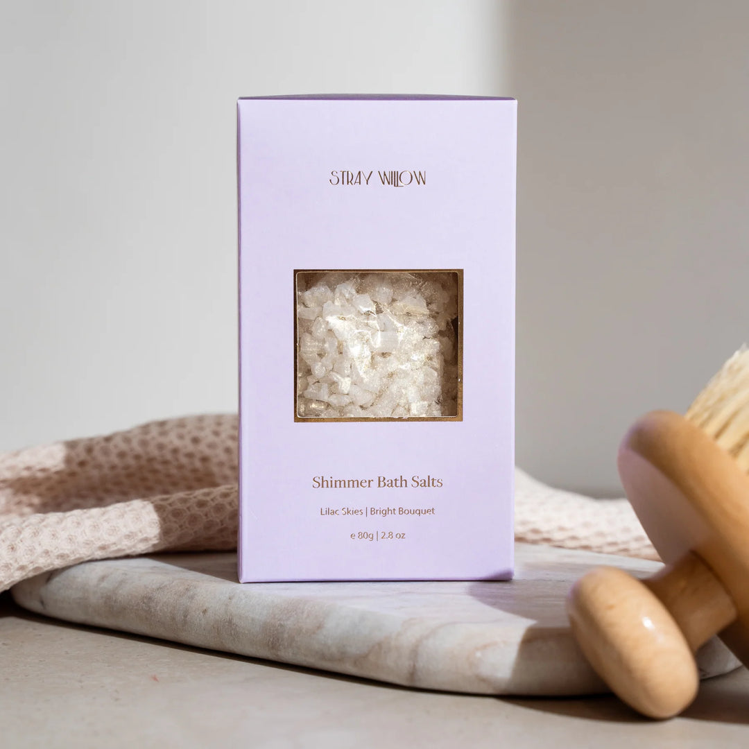 Purple box of Shimmer Bath Salts with a wooden scoop on a neutral background