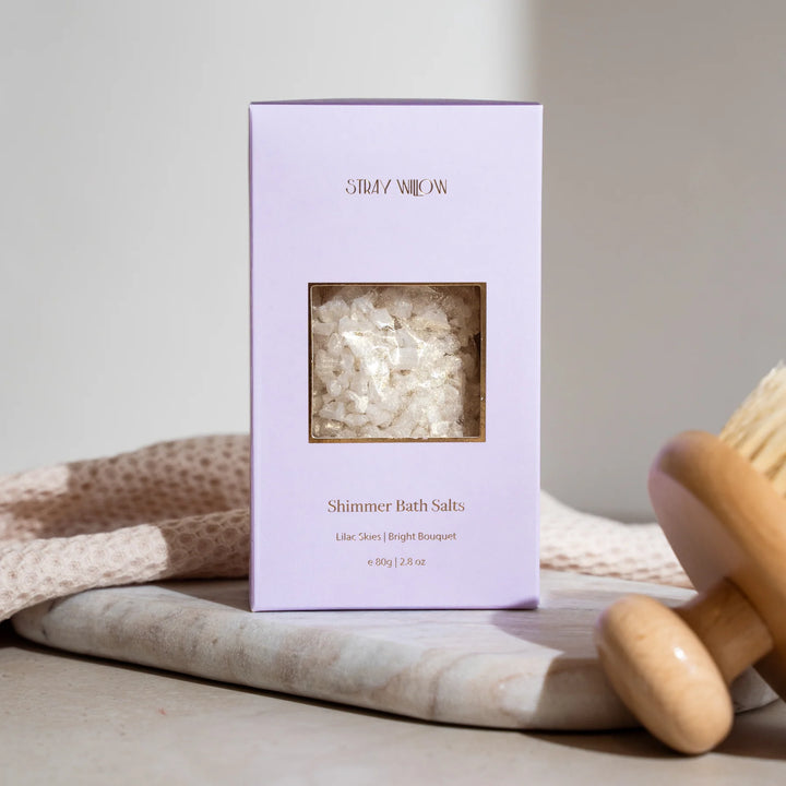 Purple box of Shimmer Bath Salts with a wooden scoop on a neutral background
