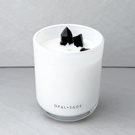 A white scented candle in a clear glass jar with three black crystal-like wicks on top, branded 'OPAL+SAGE' near the base, set against a neutral gray background.