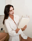 Woman in a white robe with a face mask, holding a book in a bathroom.