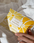 Yellow packaging with floral design held by a hand on a light fabric background