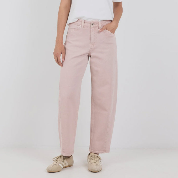 A woman modeling a pair of light pink wide-leg jeans with a white t-shirt and beige sneakers, standing against a plain light gray studio background.