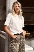 Woman wearing a white t-shirt and leopard print pants leaning against a concrete wall.