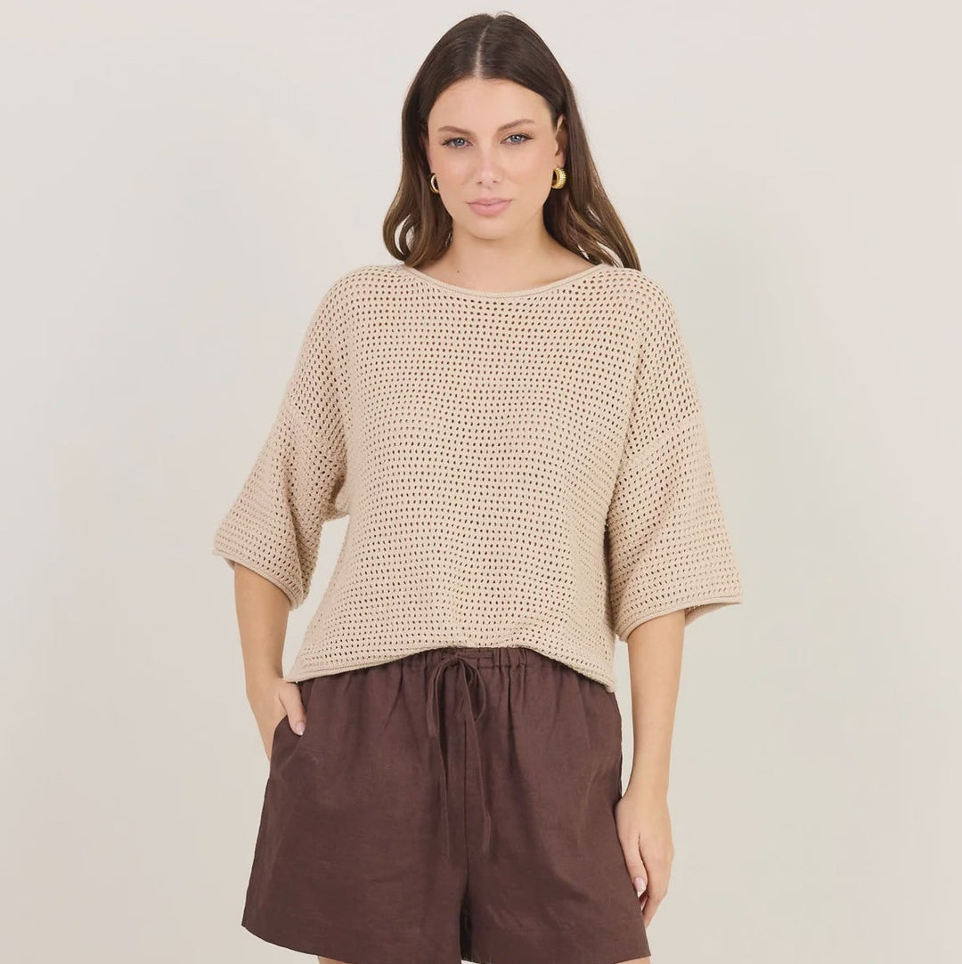 A woman modeling a beige textured knit top with elbow-length sleeves and dark brown linen shorts, paired with black flip-flop sandals, against a plain light beige studio background.
