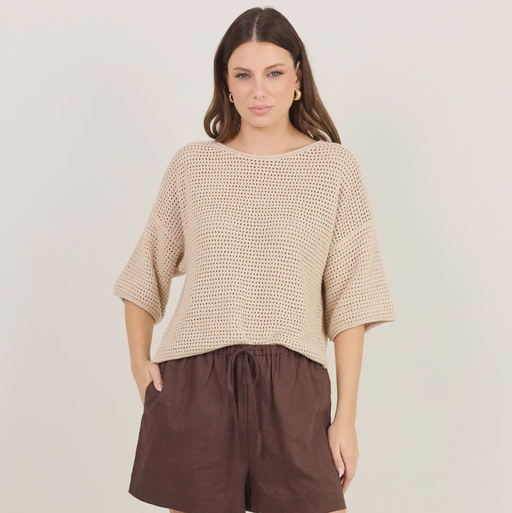 A woman modeling a beige textured knit top with elbow-length sleeves and dark brown linen shorts, paired with black flip-flop sandals, against a plain light beige studio background.