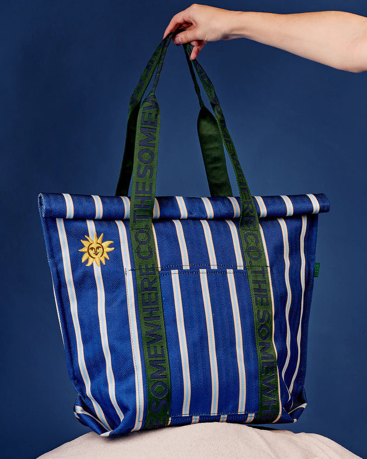 Blue and white striped tote bag with green accents and a sun design, held against a blue background.
