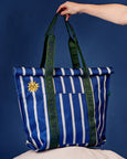 Blue and white striped tote bag with green accents and a sun design, held against a blue background.