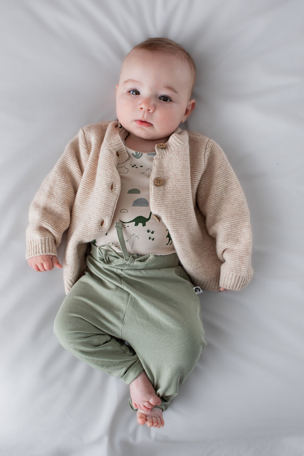 Baby wearing a beige cardigan, patterned shirt, and green pants lying on a white surface.