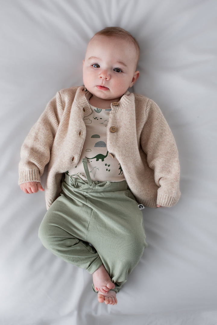 Baby wearing a beige cardigan, patterned shirt, and green pants lying on a white surface.