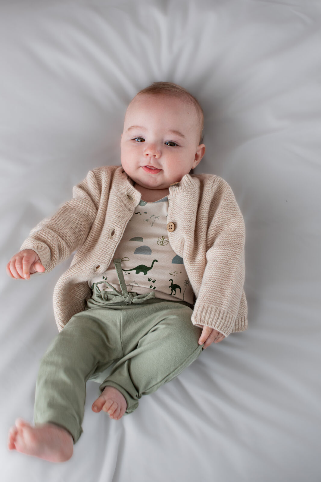 Baby wearing a beige cardigan and green pants lying on a white surface