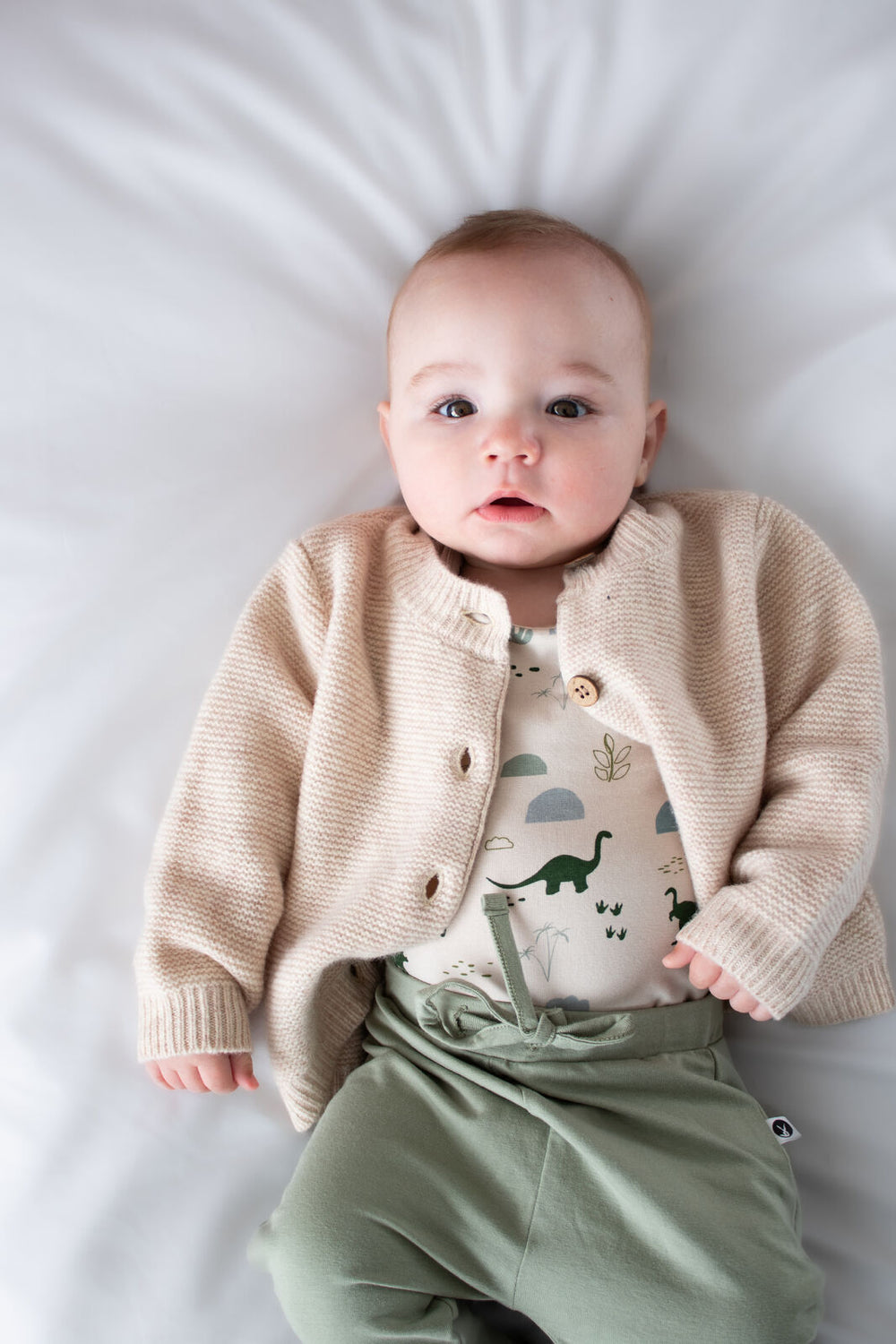 Baby wearing a beige cardigan, patterned shirt, and green pants on a white background