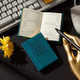 Teal password manager book with open pages on a desk next to a keyboard, mouse, and yellow flower.