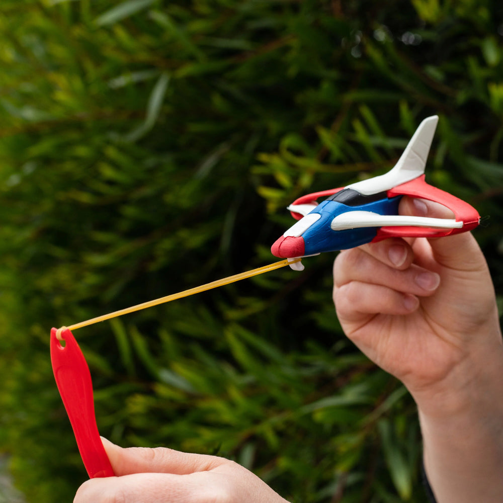 Hand holding a red, white, and blue airplane toy with a yellow string against a green leafy background