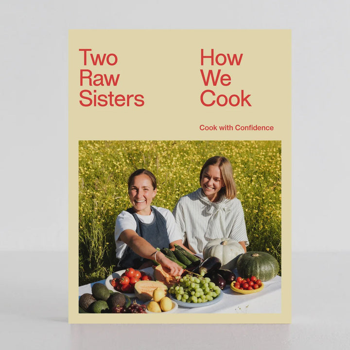 The cookbook 'Two Raw Sisters: How We Cook' stands upright on a white surface against a light gray background. The book cover features a pale yellow background with red text and a central color photograph of two women outdoors among green plants and fresh produce.