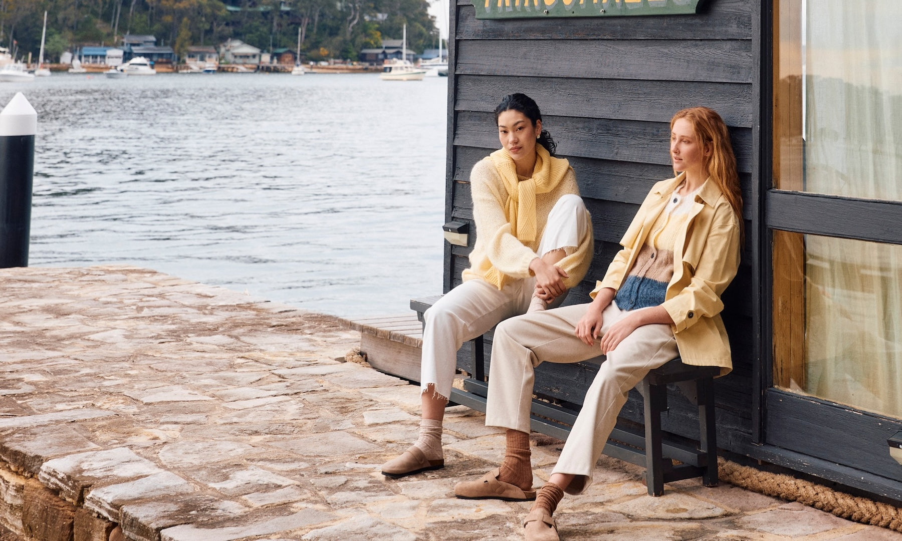 Two women sitting on a dock by a body of water, wearing matching yellow jackets.
