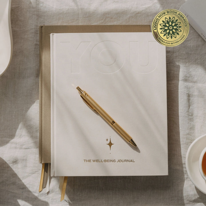 A white hardcover journal titled 'YOU' with 'THE WELLBEING JOURNAL' printed on the cover, styled in a flat lay with a gold pen, a gold seal sticker, and surrounding tea, ceramic dishes, and a magazine on a linen surface.
