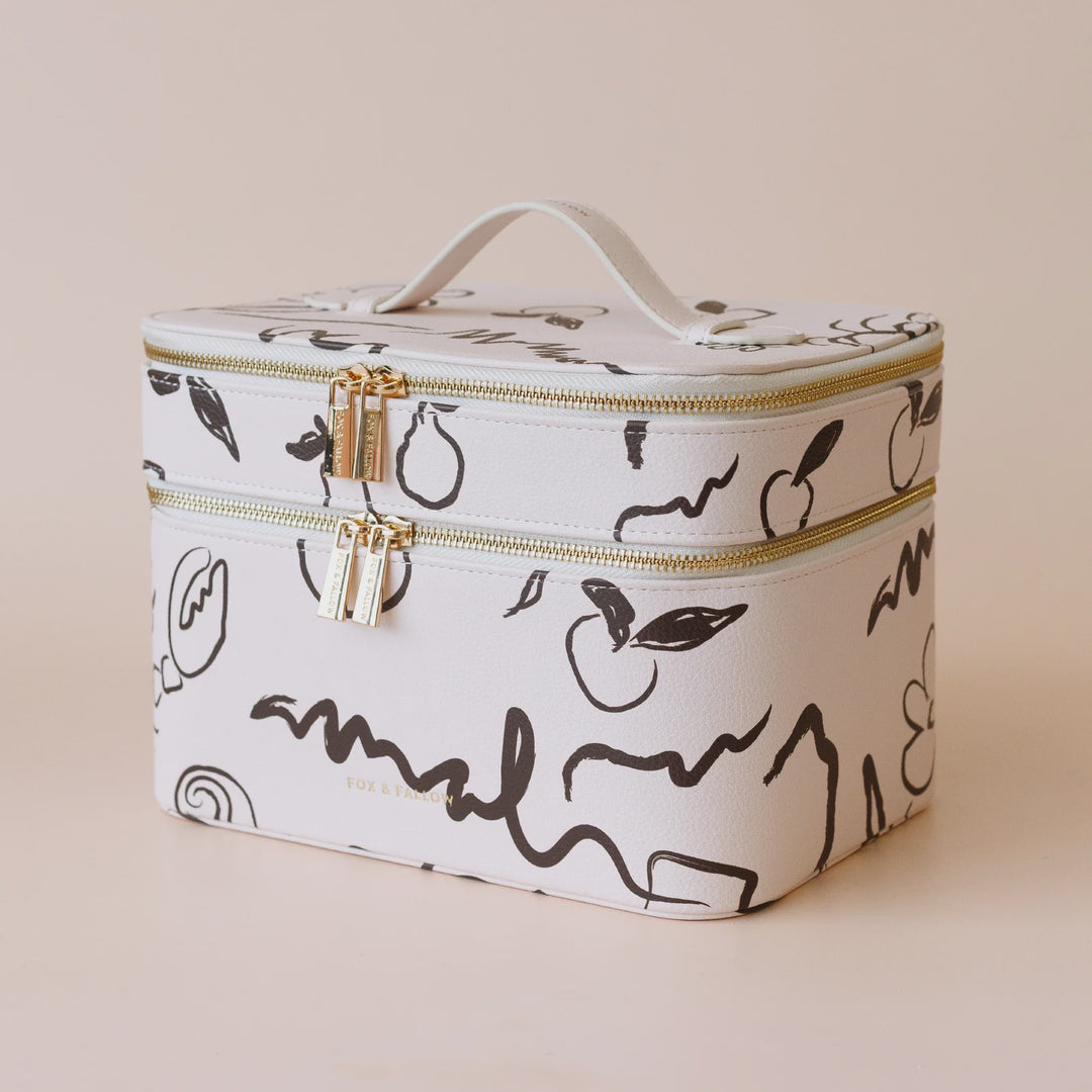 A white cosmetic case with a black fruit and leaf pattern, featuring gold-tone zippers and a top handle, set against a solid beige background.