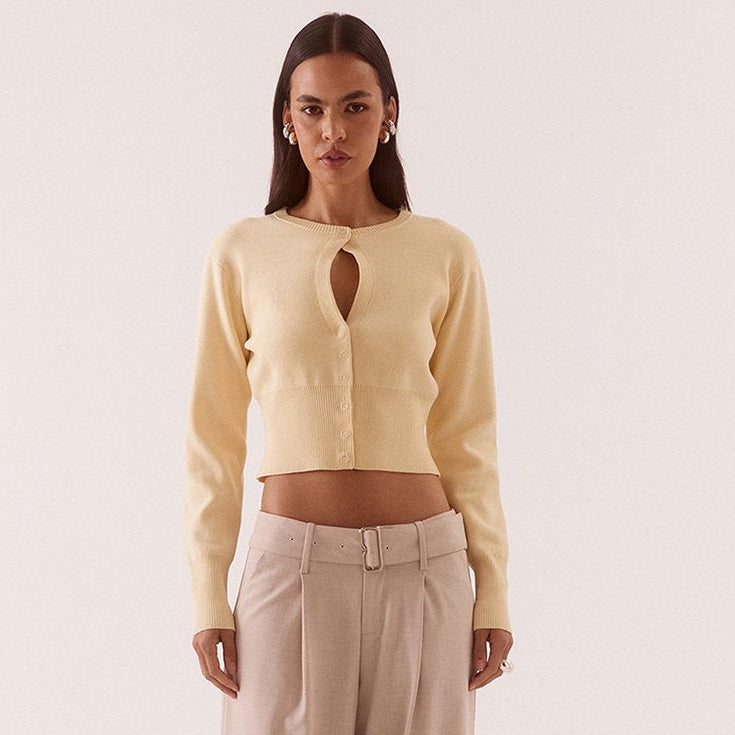 A model stands against a plain light beige studio background wearing a pale yellow cropped cardigan with a keyhole cutout and beige wide-leg trousers.