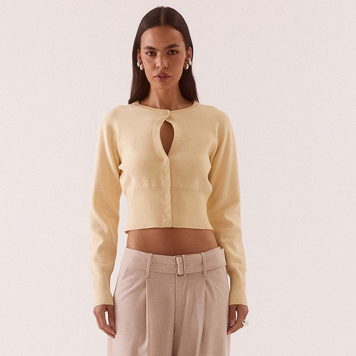 A model stands against a plain light beige studio background wearing a pale yellow cropped cardigan with a keyhole cutout and beige wide-leg trousers.